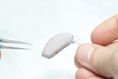 Audiologist Performs Hearing Aid Servicing image is a closeup showing fingertips holding a hearing aid on the right while a pair of tweezers is in the frame on the left. 
