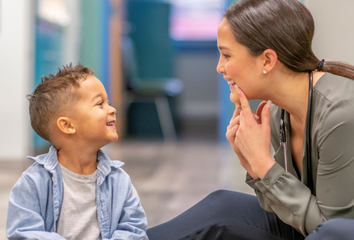 female speech therapist smiling at a small child and pointing at the corners of her mouth as the the child watches and smiles back