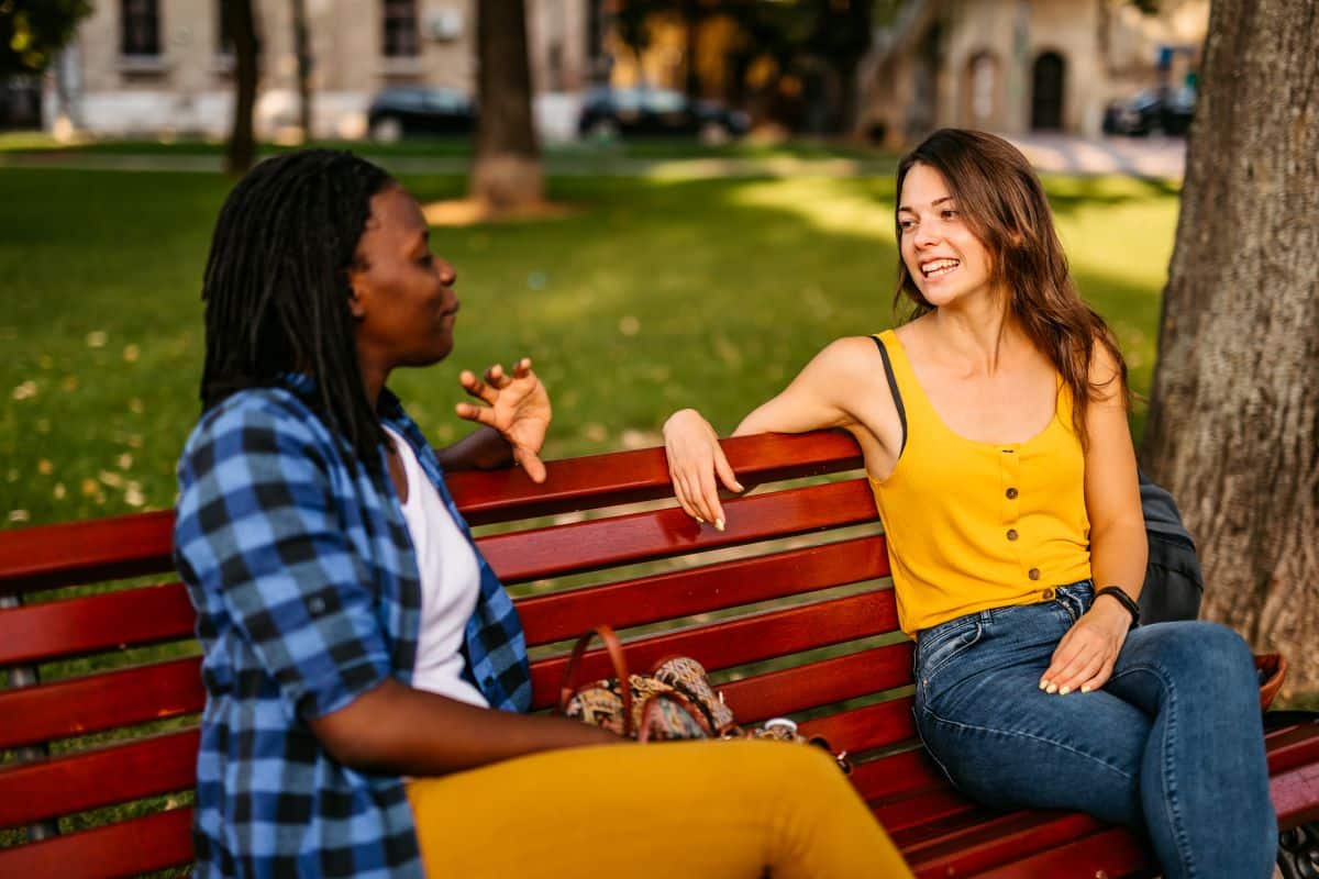 two women talking on a red park bench, one is black and wearing a blue shirt and yellow pants, the other is white and wearing a yellow shirt and blue pants