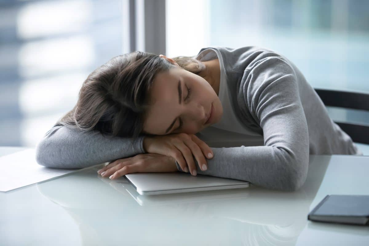 Young woman who has fallen asleep on an office desk with a closed laptop underneath her. She is resting her head on her crossed arms. 