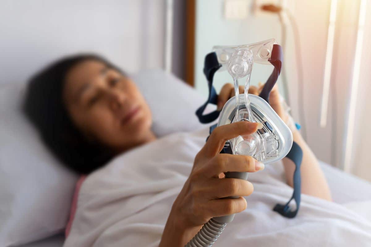 Woman in a hospital clinical setting is laying on a hospital bed, she is blurry and the focus of the image is on the half-mask for a cpap that she is holding up in front of herself. 