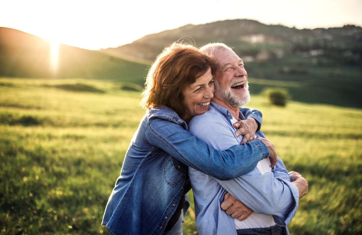 Woman and man in a field with sun going down. Woman is holding man from behind in a familiar way as he laughs. 